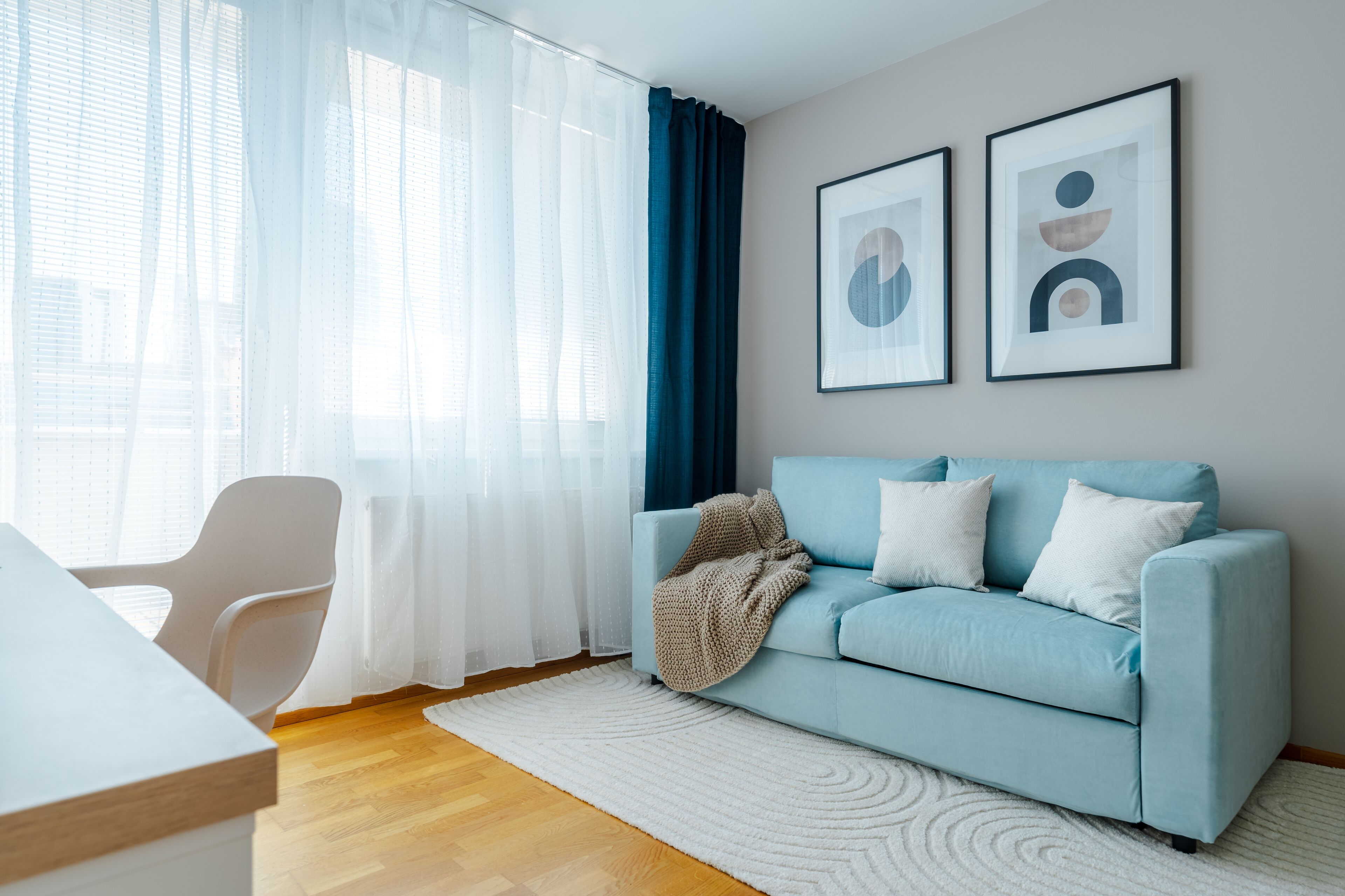 A modern living room with a light blue sofa, white cushions, a white rug, a white chair, and two framed abstract art pieces on the wall.