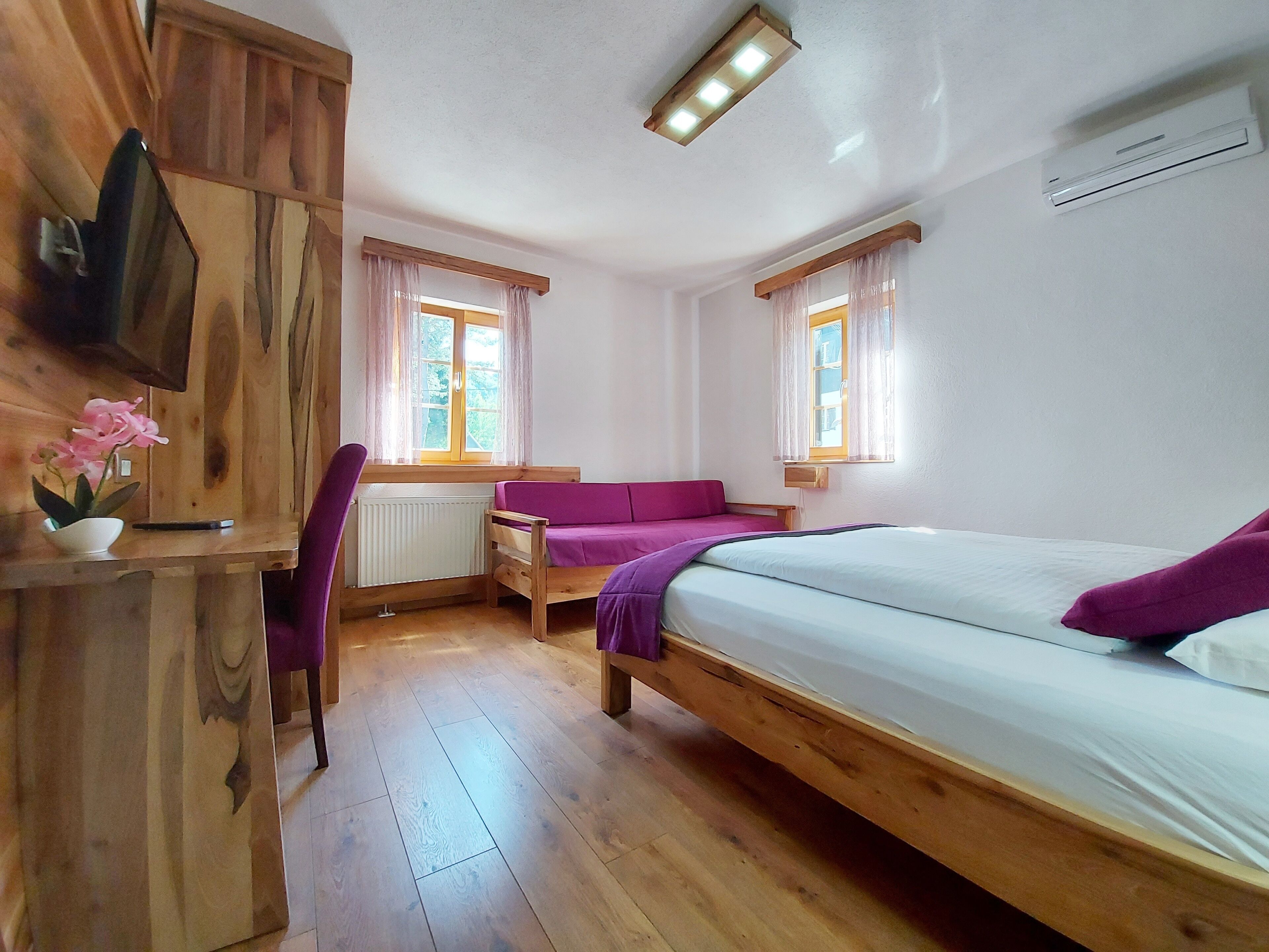 Ein gemütliches Zimmer mit einem Holzbett, einem Schreibtisch und einem Stuhl. Zwei Fenster mit Vorhängen, ein Heizkörper und ein an der Wand befestigtes Fernsehgerät sind vorhanden.