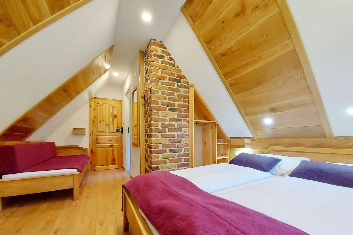 A bedroom with a sloped ceiling, wooden walls, a brick fireplace, a bed with a purple blanket, and a wooden bench.