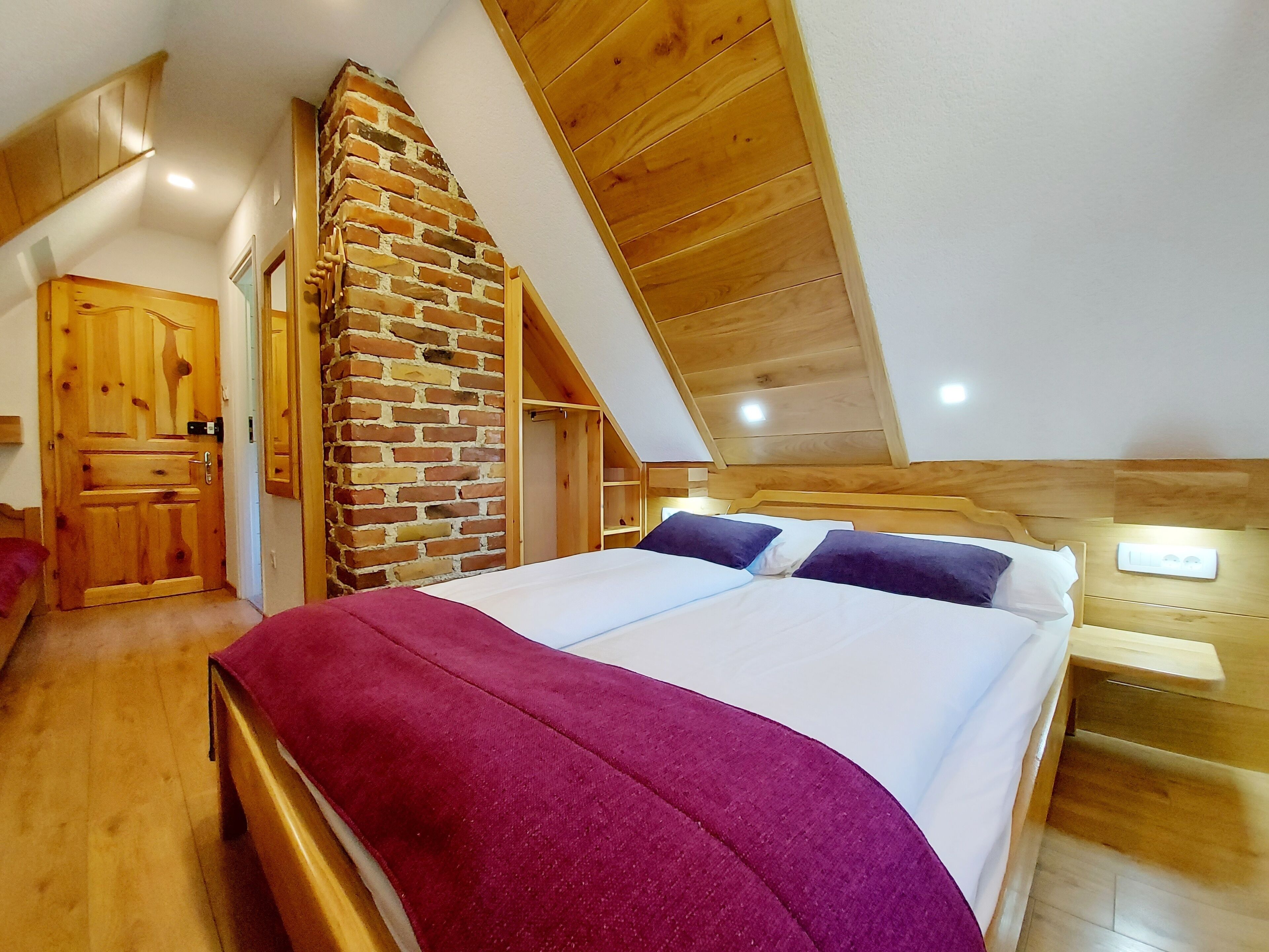 Ein Schlafzimmer mit geneigter Decke, einem Backstein-Kamin, Holzwänden und einem Bett mit violetter Decke.