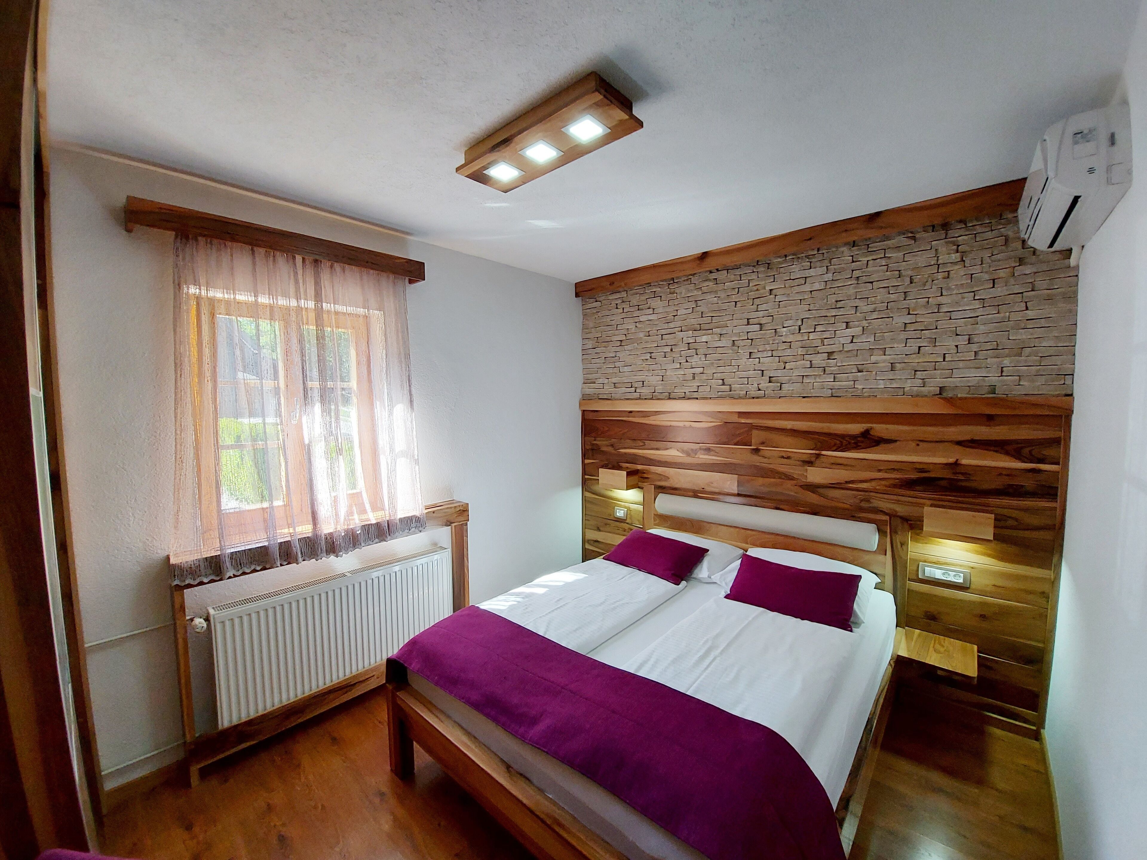 Une chambre avec une tête de lit en bois, un radiateur blanc, une fenêtre avec des rideaux légers et un mur décoré d’un motif en briques.
