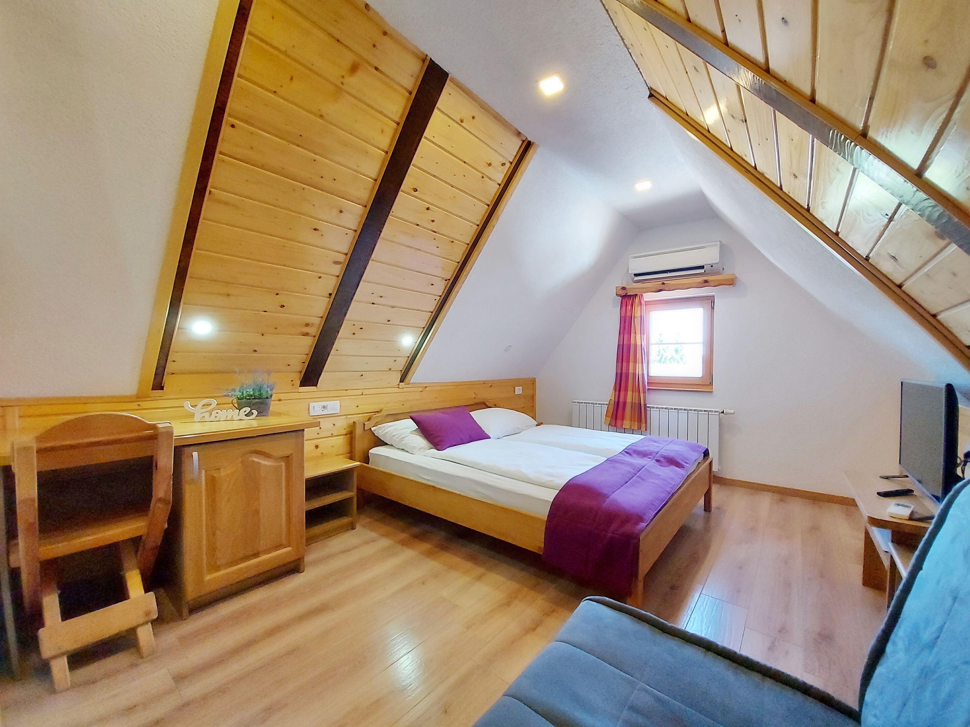 A cozy attic bedroom with a sloped ceiling, wooden walls, a bed with a purple blanket, a desk, a chair, and a TV.