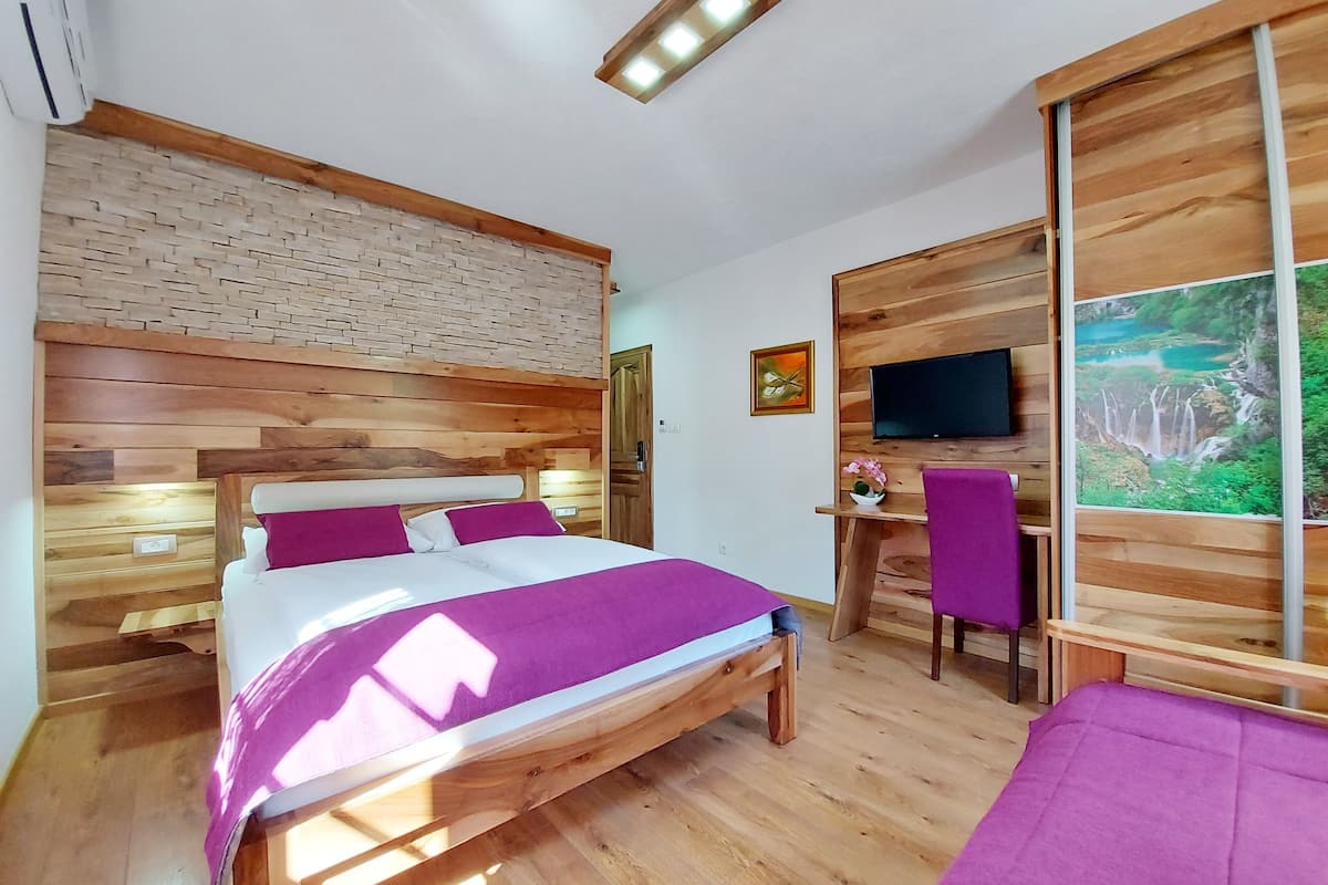 A bedroom with a wooden headboard, a bed with a purple bedspread, a desk with a chair, a television, and a large window with a view of a waterfall.