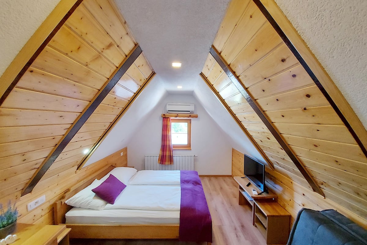 A small, wooden-paneled attic bedroom with a bed, a TV, and a window with a curtain.