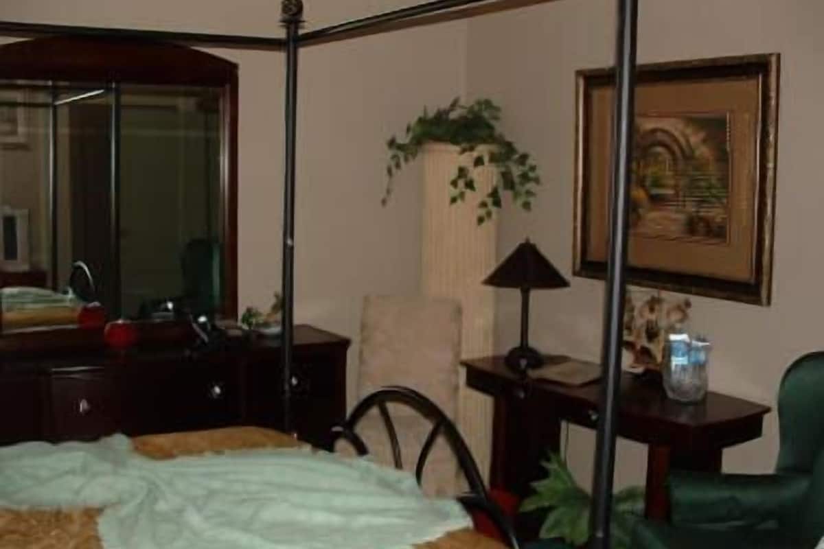 A bedroom with a four-poster bed, a desk with a lamp, a chair, a mirror, and a framed picture on the wall.