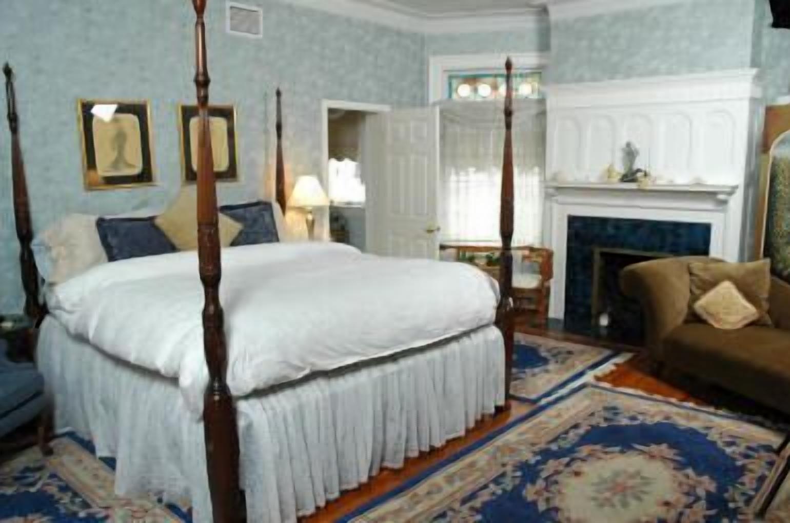 A four-poster bed with white linens, a brown sofa, a fireplace, and a patterned carpet.