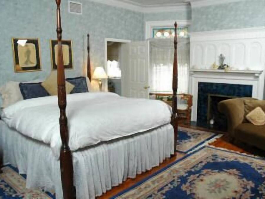 A four-poster bed with white linens, a brown sofa, a fireplace, and a patterned carpet.