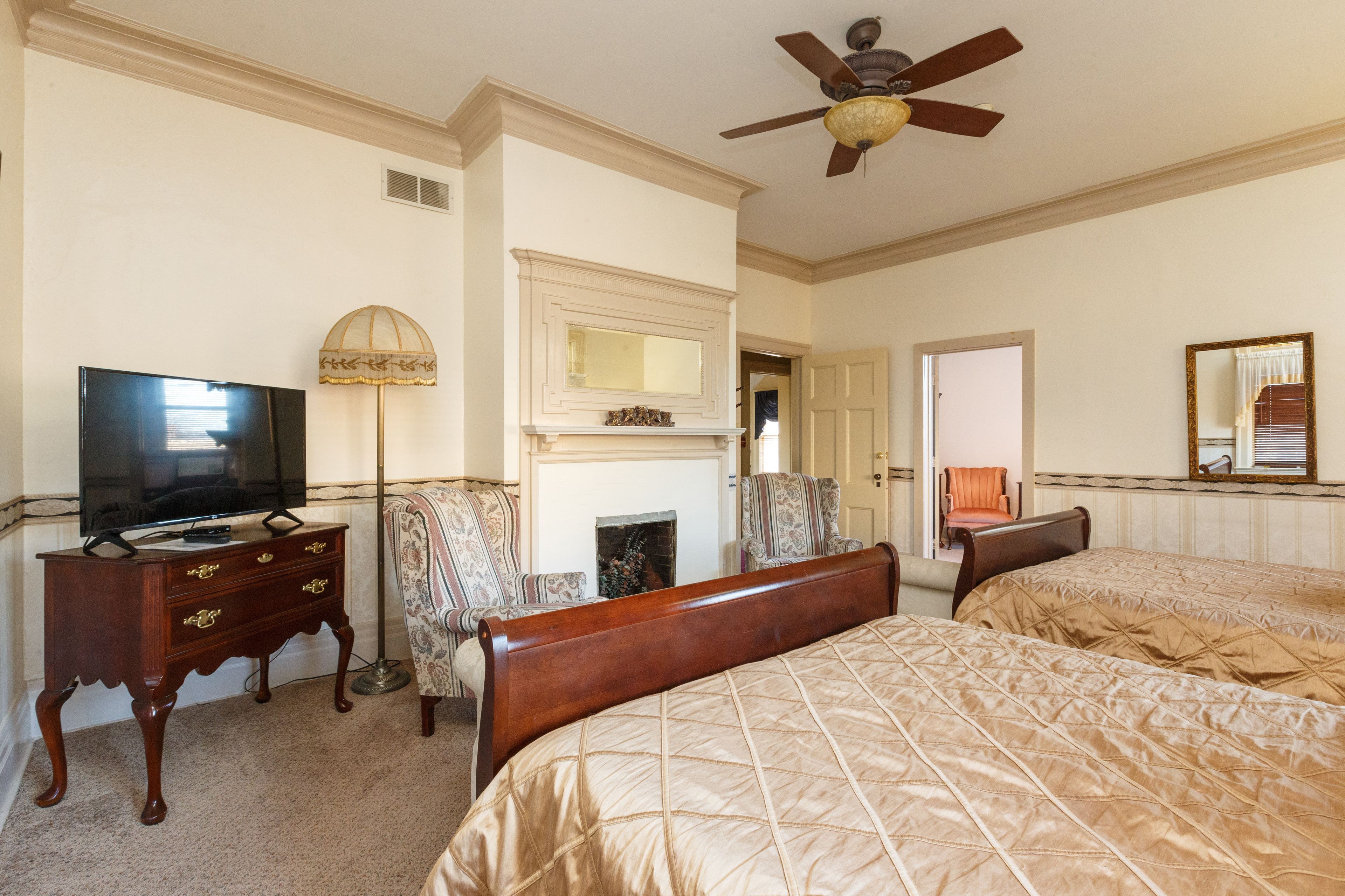 A hotel room with a television, a fireplace, two beds, a ceiling fan, and a mirror.