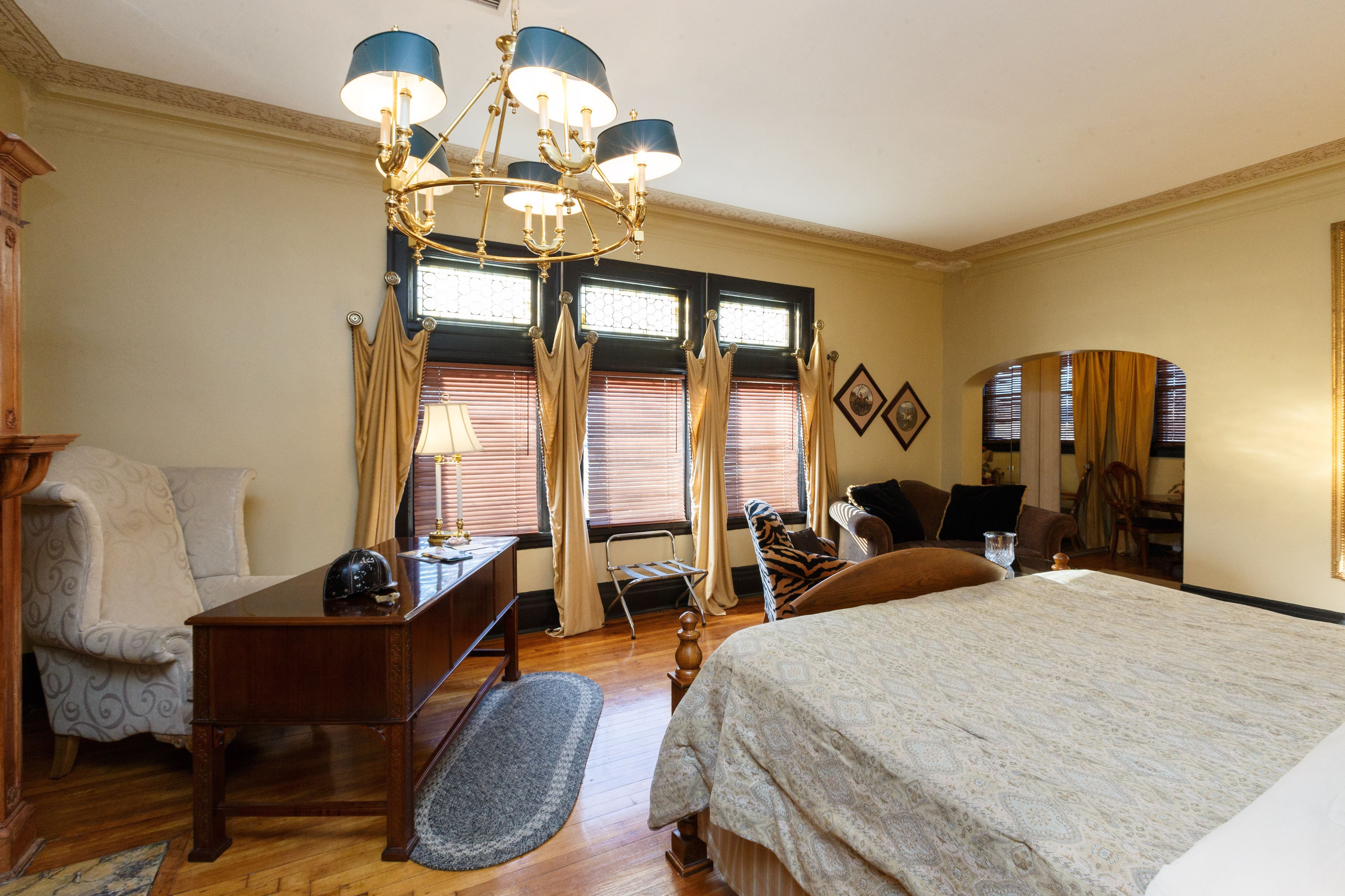 A hotel room with a large bed, a desk with a lamp, a chair, a small table, and a window with curtains.