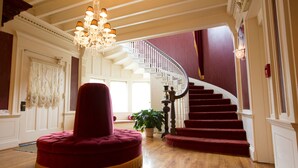A grand staircase with a red carpet, a chandelier, and a red cushioned ottoman in a spacious room with wooden flooring. - Boggs Mansion (Pittsburgh)
