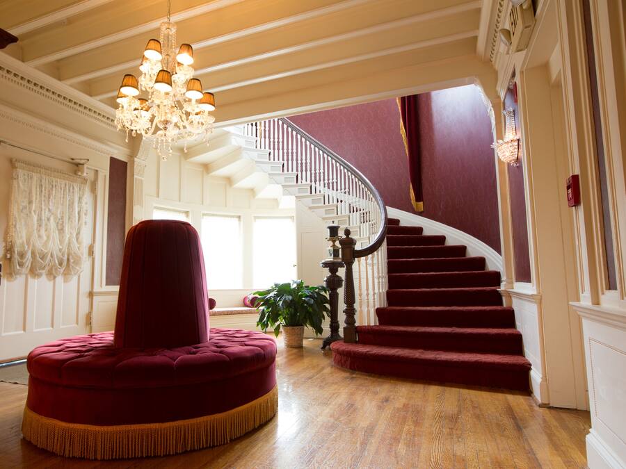 Una gran escalera con alfombra roja, un candelabro y un otomán tapizado en rojo en una amplia habitación con piso de madera.