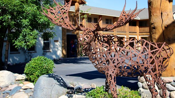 A metal sculpture of a moose in front of a building with a wooden facade and a balcony. - Moose Creek Lodge & Suites (Cody)