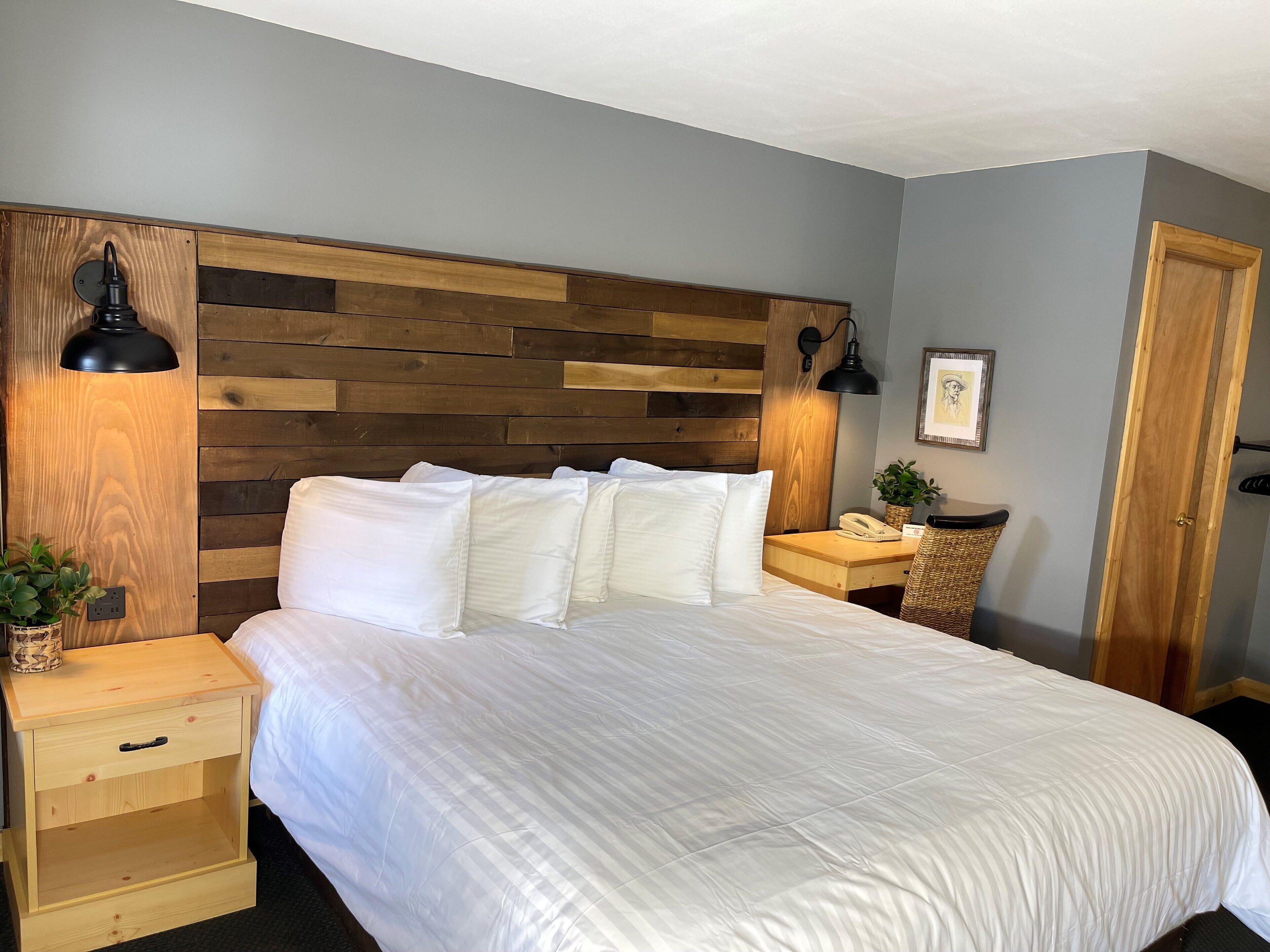 Un dormitorio con cabecera de madera, cama con ropa de cama blanca, dos mesitas de noche con lámparas, una mesita auxiliar, una planta pequeña y una puerta.