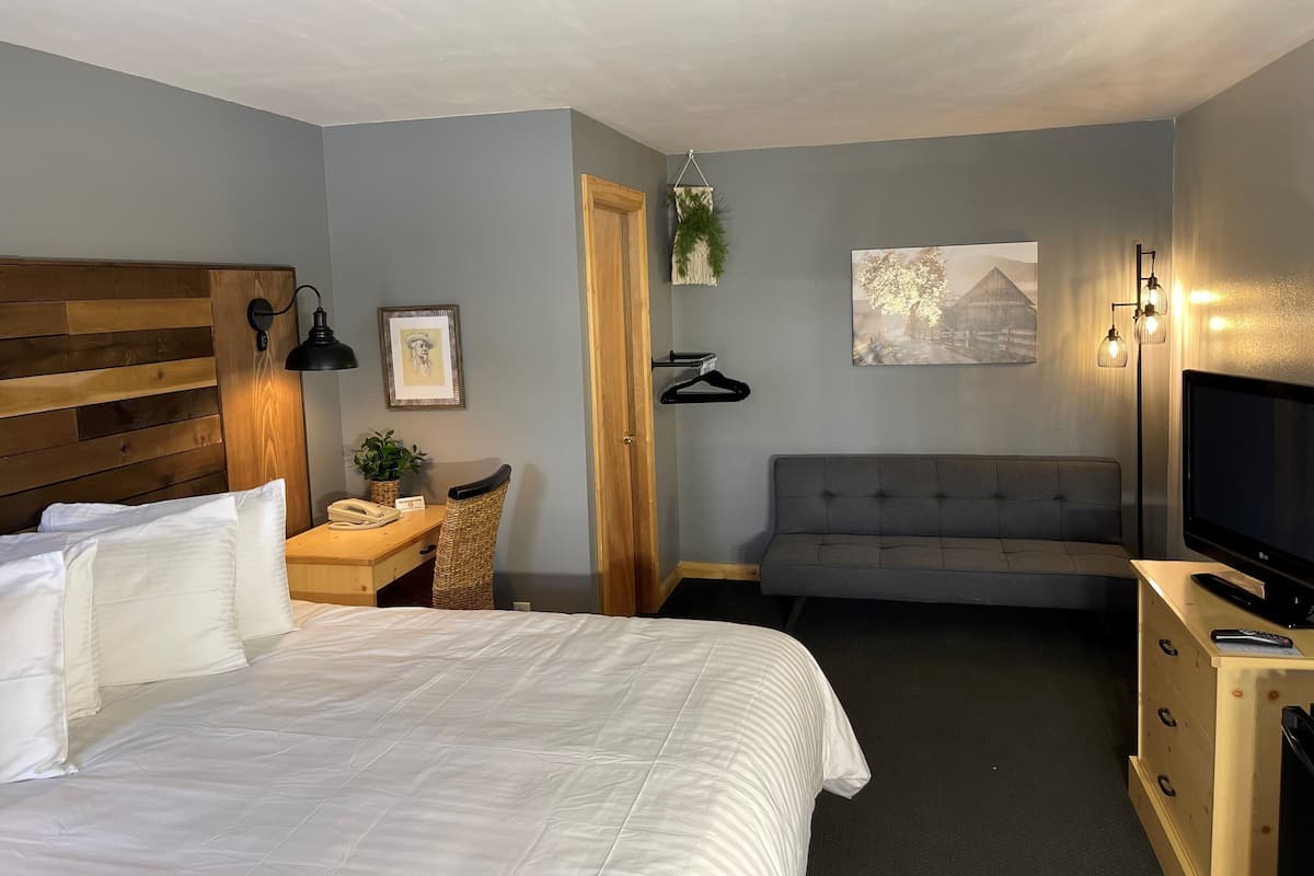 A hotel room with a bed, bedside table, lamp, chair, desk, TV, and a sofa.