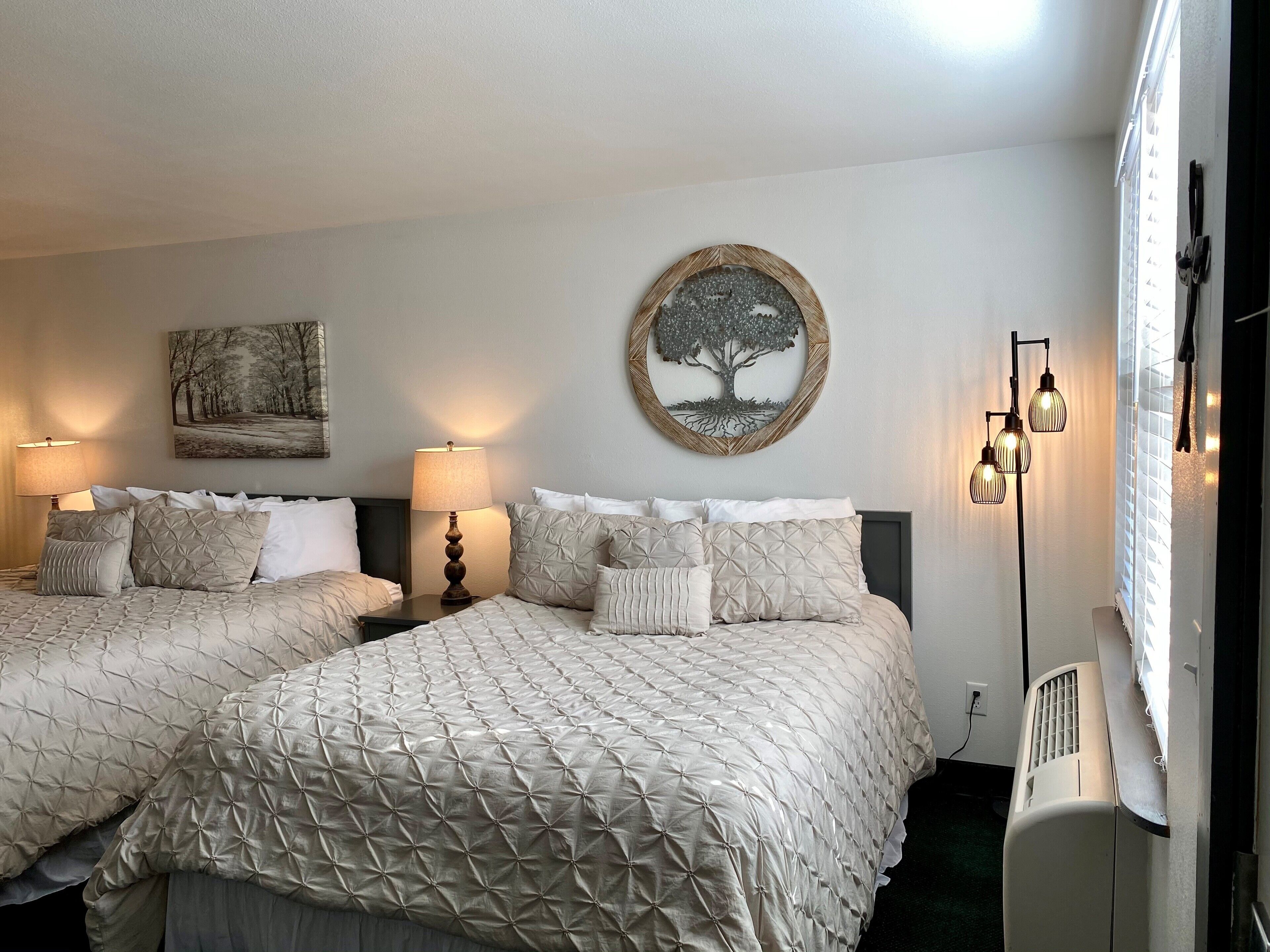 Habitación de hotel con dos camas, una obra de arte de árbol colgada en la pared, una lámpara y un ventilador de techo.