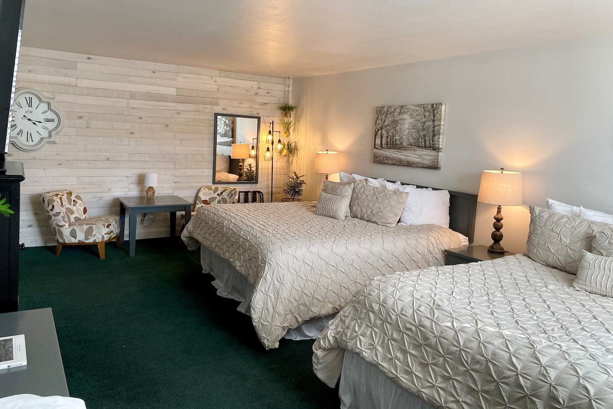 A hotel room with two beds, a wooden wall, a desk with a chair, a clock, and a mirror.