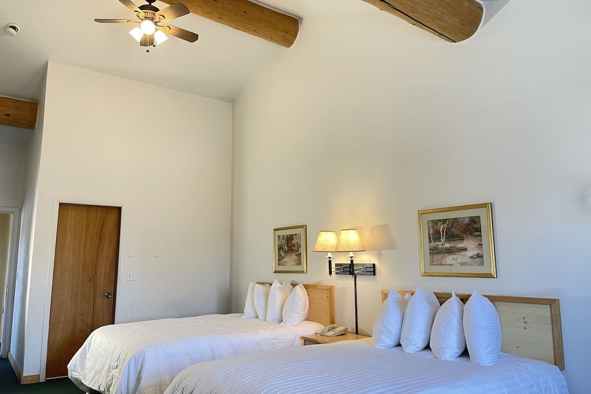 A hotel room with two beds, a ceiling fan, a wooden door, and two wall-mounted lamps.