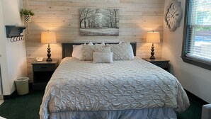 A neatly arranged bedroom with a large bed, two bedside tables, a wall clock, and a window with blinds. - Moose Creek Lodge & Suites (Cody)