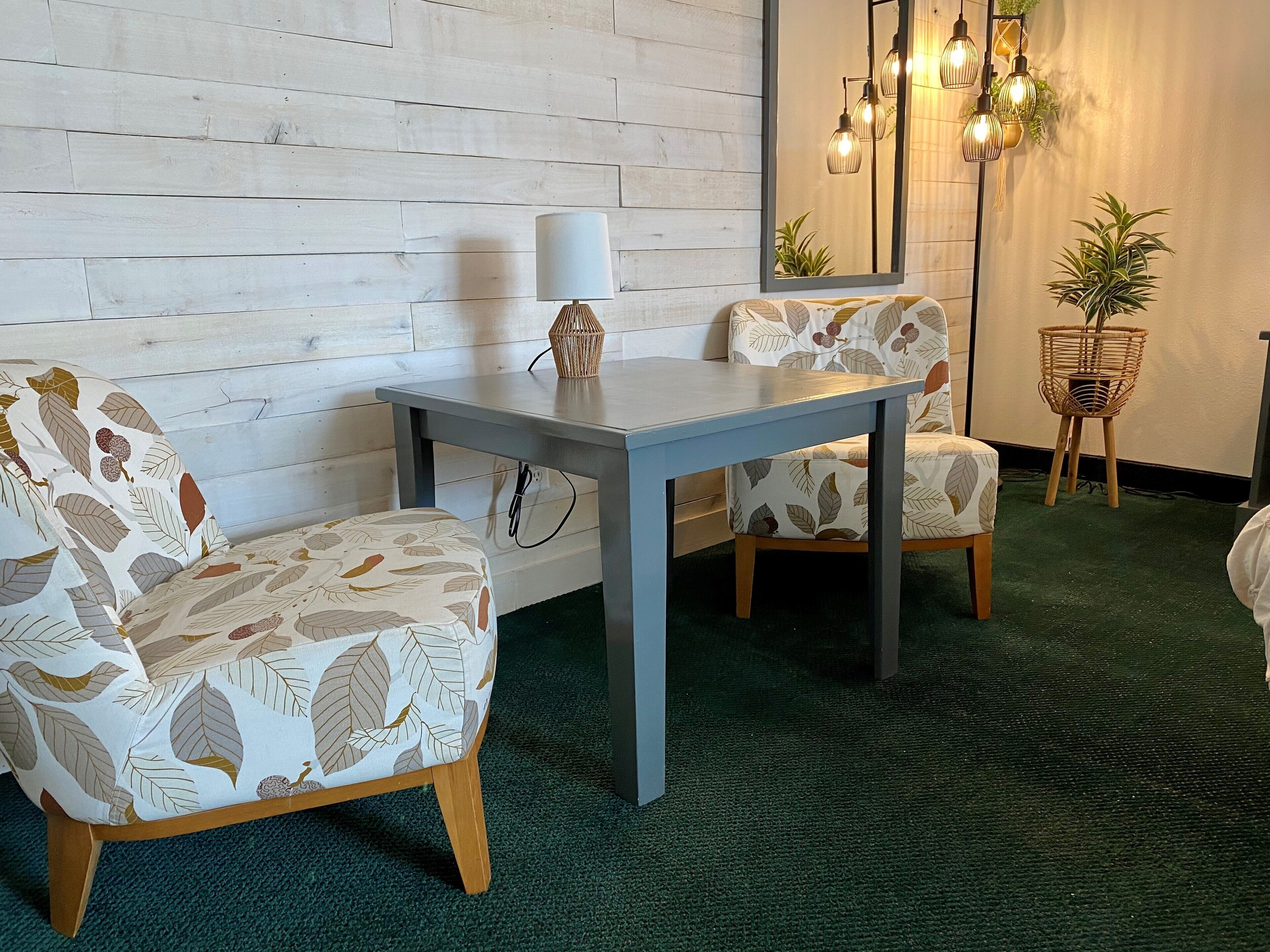A room with a wooden wall, a grey table, two patterned chairs, a lamp, a mirror, and hanging lights.