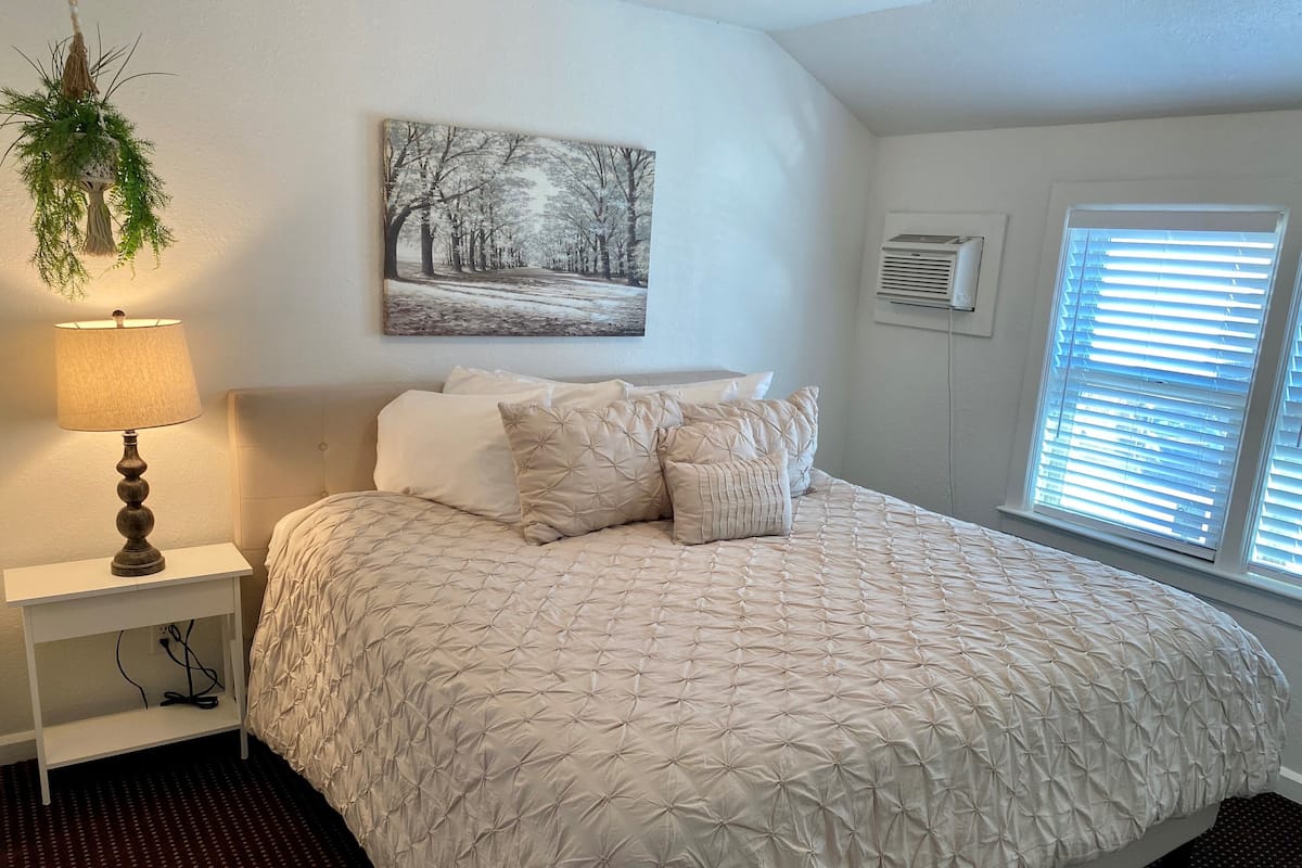 A bedroom with a bed, a bedside lamp, a wall-mounted air conditioner, and a window with blinds.