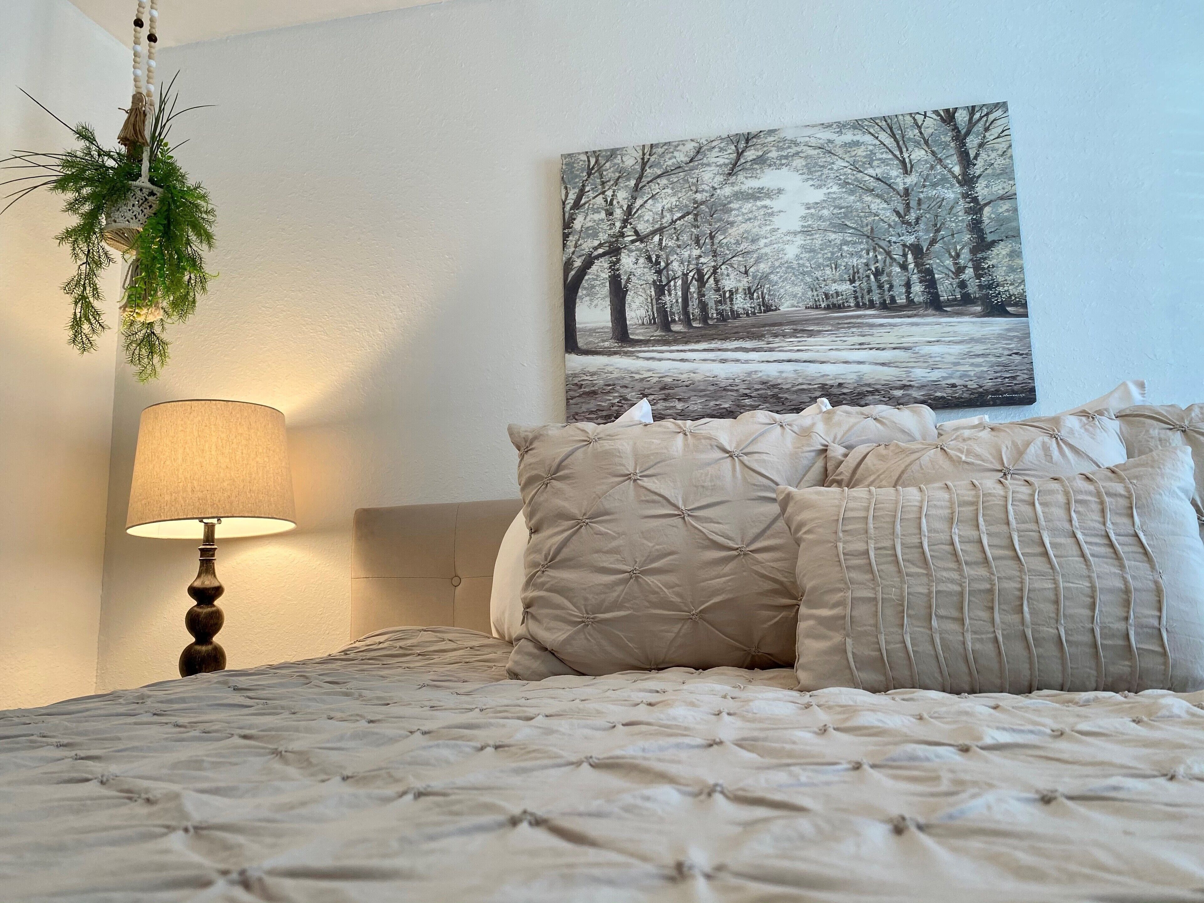 Et soveværelse med en seng, en lampe, en plantekasse på væggen og et billede af en skov.