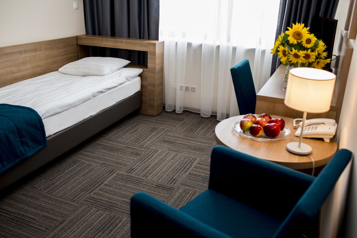 A hotel room with a single bed, a small table with a lamp, a chair, and a vase of sunflowers.