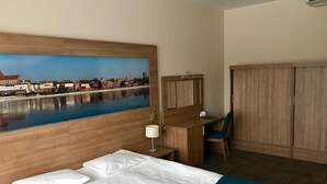 A hotel room with a bed, a desk, a chair, a wardrobe, and a large window with a cityscape view. - Hotel Meeting (Torun)