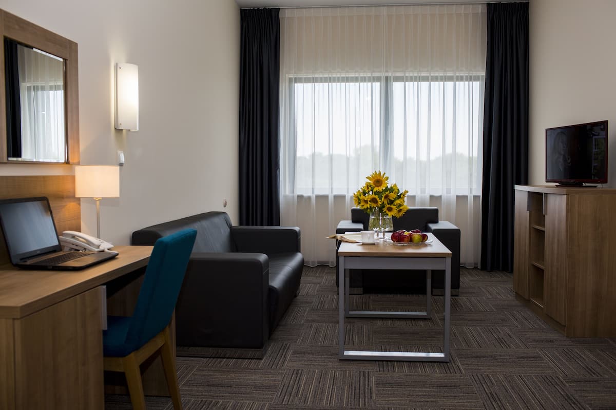 A hotel room with a desk, a laptop, a telephone, a sofa, a coffee table with a vase of sunflowers, a television, and a wardrobe.