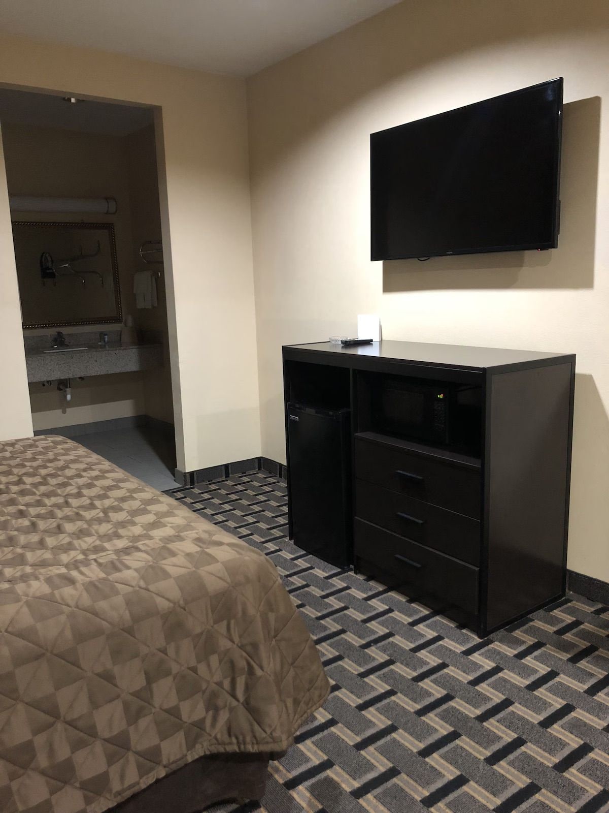 Ein Hotelzimmer mit Bett, Schreibtisch mit Schubladen, einem an der Wand befestigten Flachbildfernseher und einem Badezimmer mit Spiegel und Waschbecken.