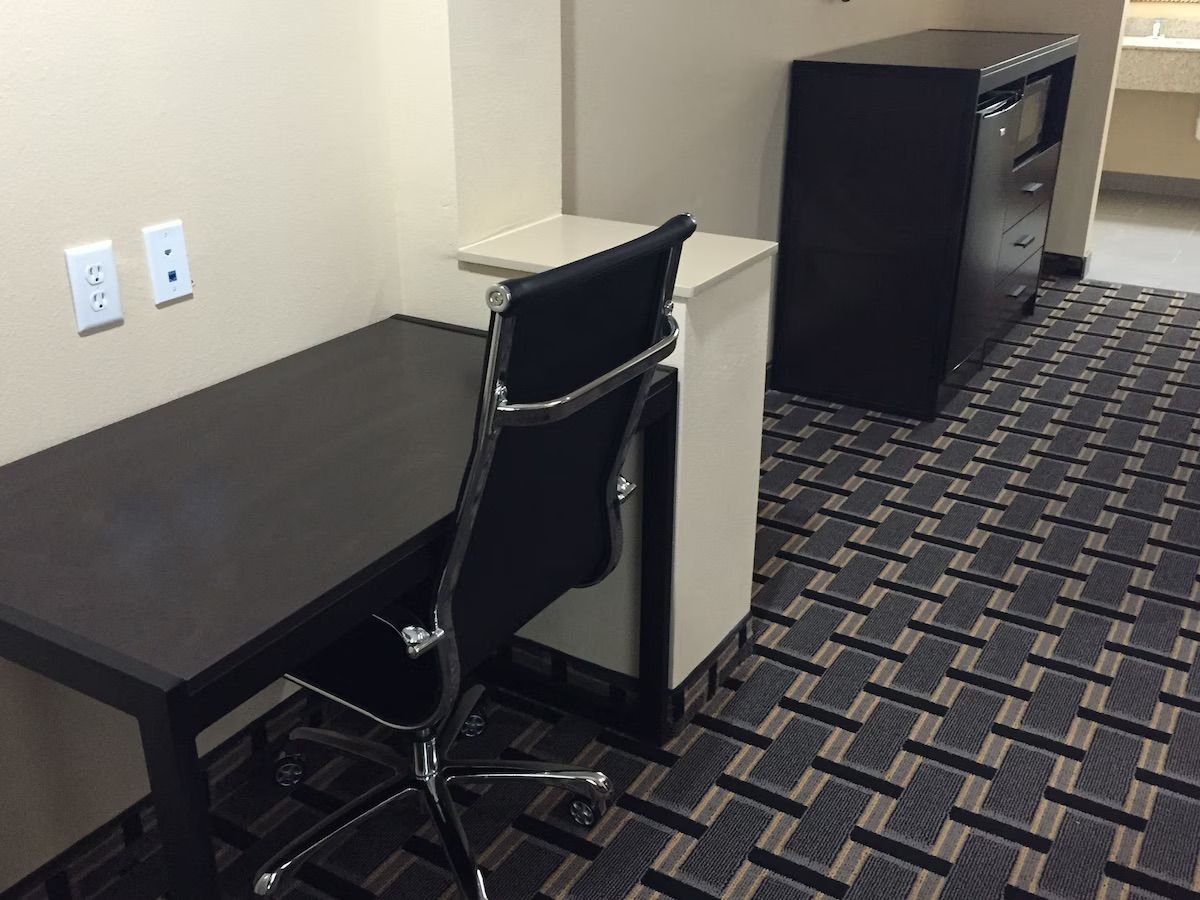 Una habitación de hotel con un escritorio negro, una silla de oficina negra, un armario blanco y un archivador negro.