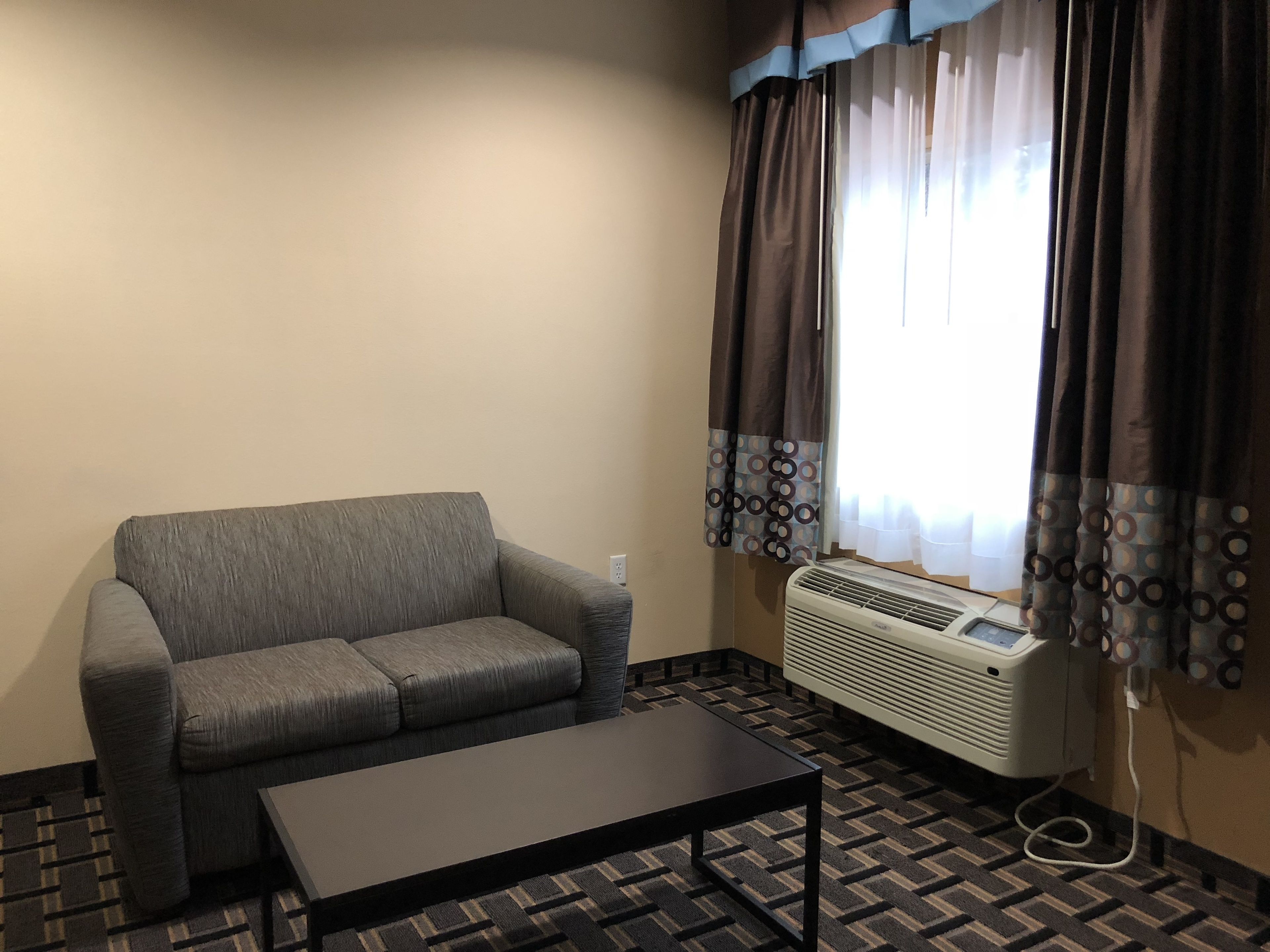 Una habitación pequeña amoblada con un sofá gris, una mesa de centro negra y una ventana con cortinas.