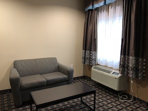 Une petite pièce meublée, avec un canapé gris, une table basse noire et une fenêtre avec des rideaux.
