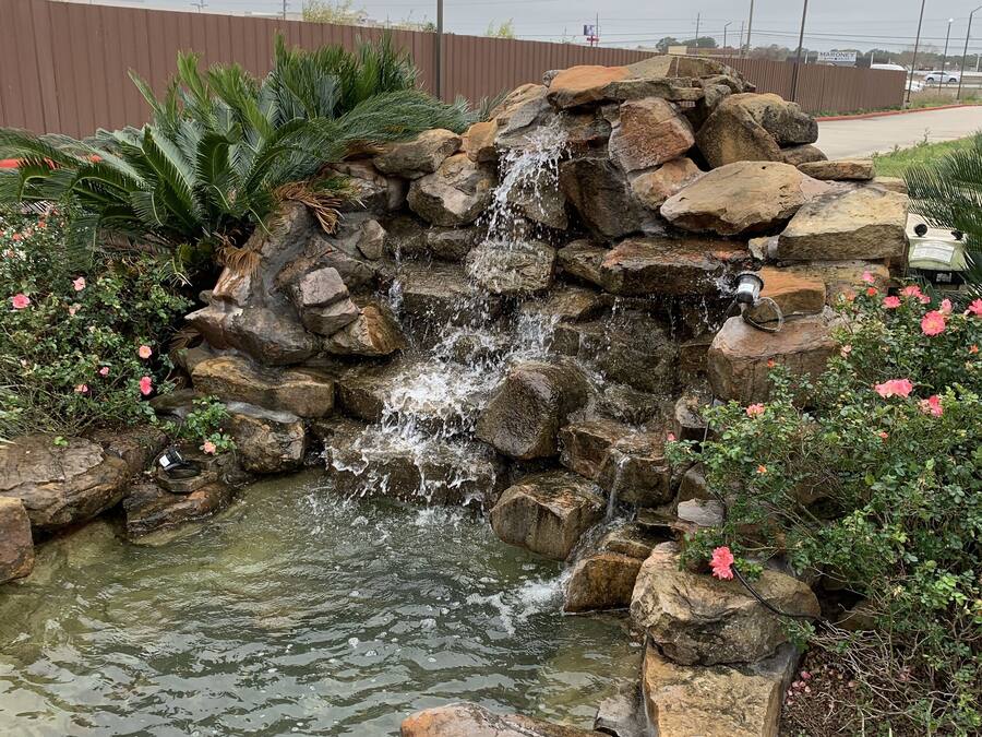 Una pequeña cascada rodeada de rocas y plantas, con una zona pavimentada al fondo.