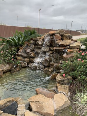 Petita cascada enmig de roques i vegetació, amb una zona pavimentada al fons.