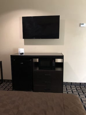 A compact hotel room with a bed, a desk with a flat-screen TV, a microwave, and a small refrigerator. - Downtowner Inn (Houston)