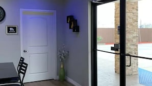 A room with a white door, a small table with a chair, a vase with flowers, and a view of an outdoor area with a blue mat and a stone pillar.
