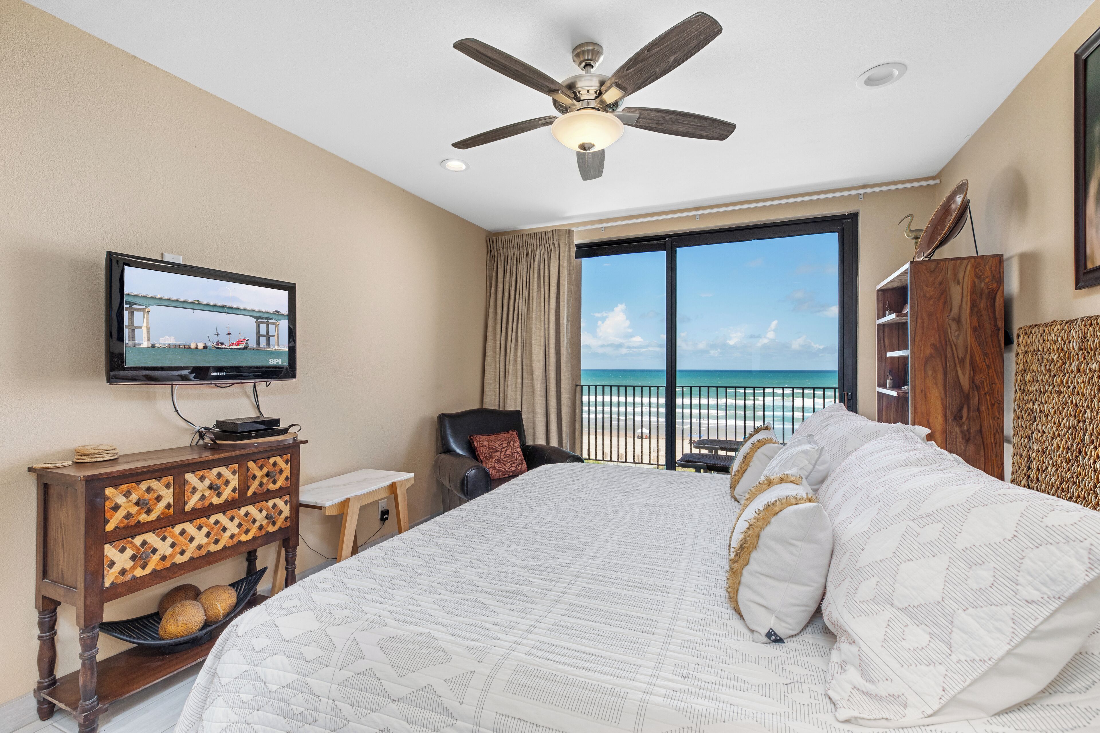 Una camera d'albergo con un letto grande, una ventola a soffitto, una televisione su un mobile in legno, vista sulla spiaggia e un balcone con tavolo e sedia.