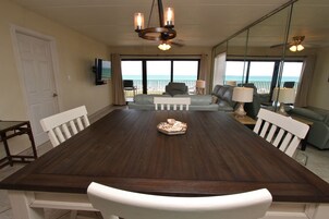 Un comedor con una gran mesa de madera, sillas blancas, un sofá y un televisor en la pared.
