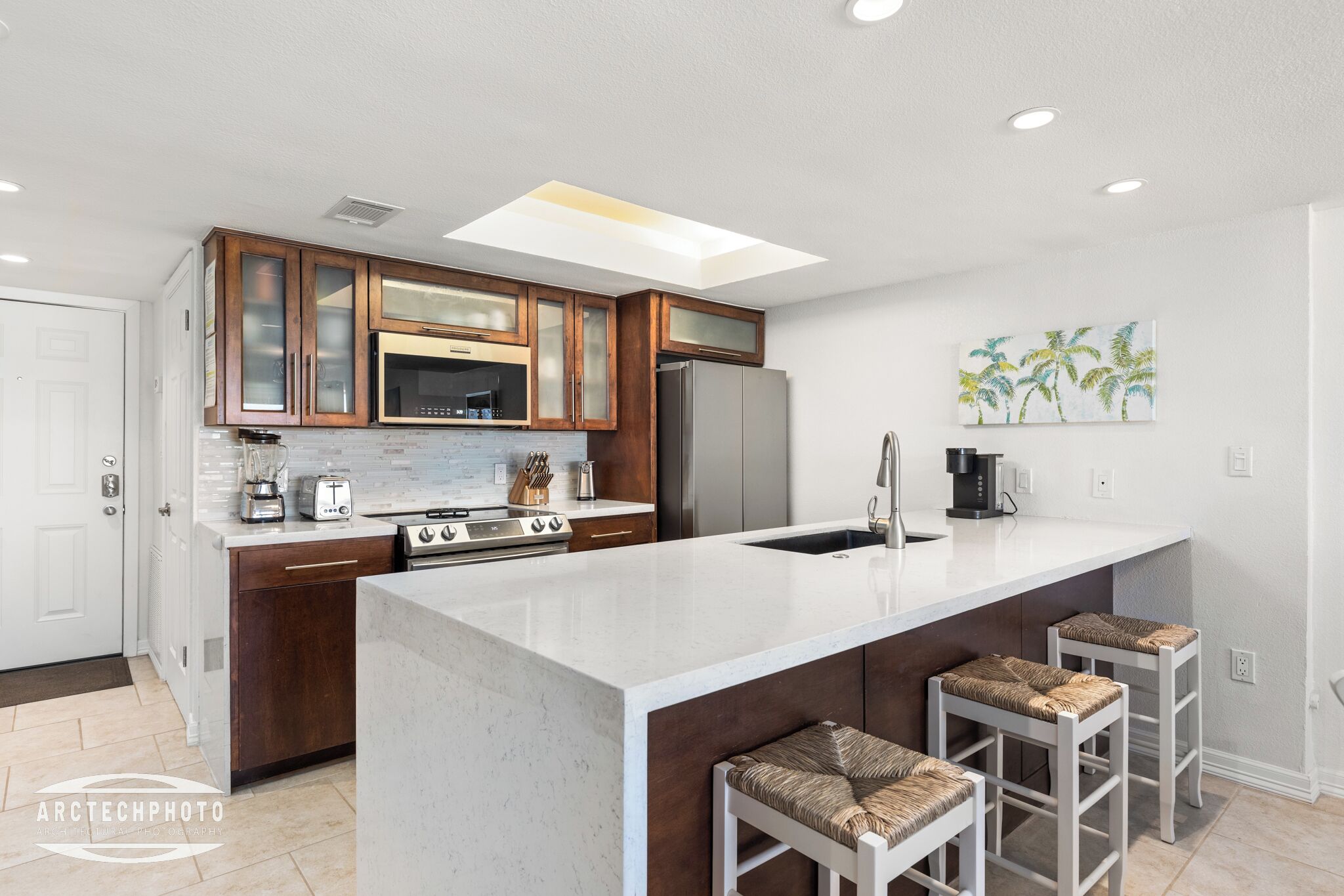 A modern kitchen with wooden cabinets, a central island with stools, a microwave, a refrigerator, and a coffee maker.