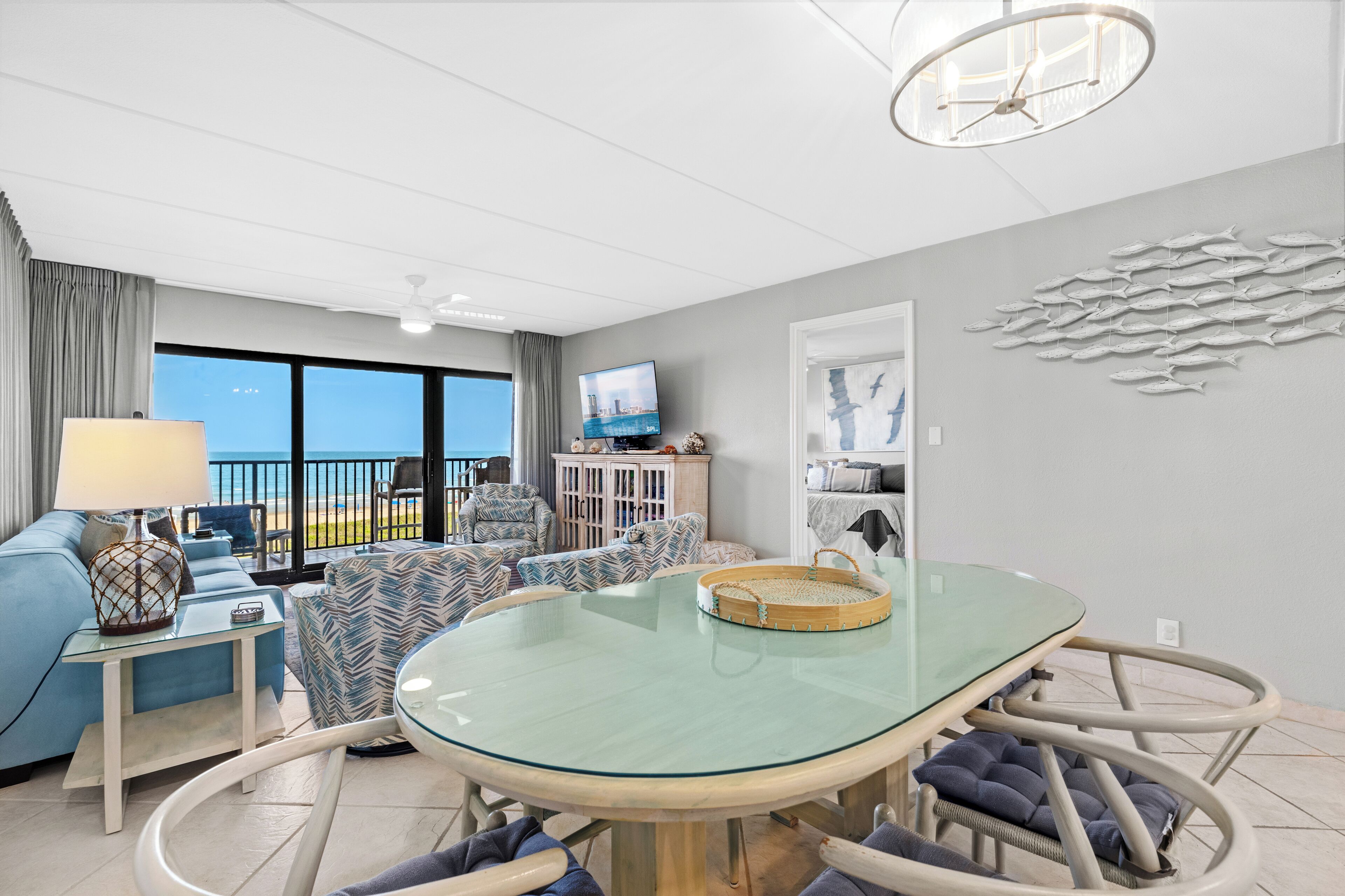 Een moderne woonkamer met een tafel met glazen blad, blauwe en gestreepte banken, een televisie en uitzicht op zee.