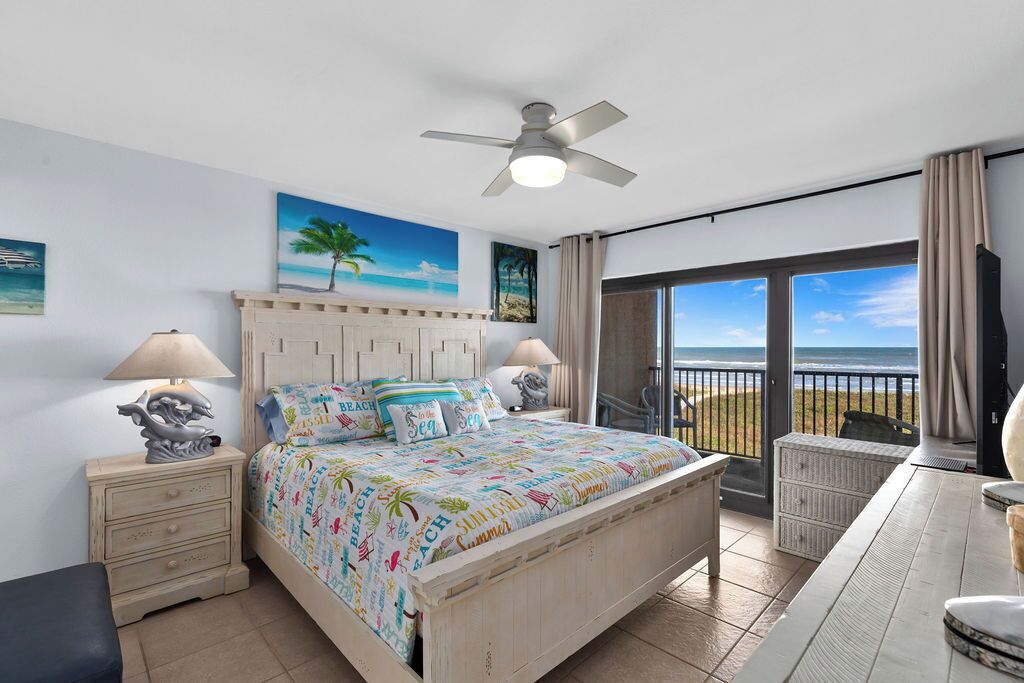 Un dormitorio con cama, mesita de noche, lámpara, ventilador de techo y vista a la playa a través de un ventanal grande.
