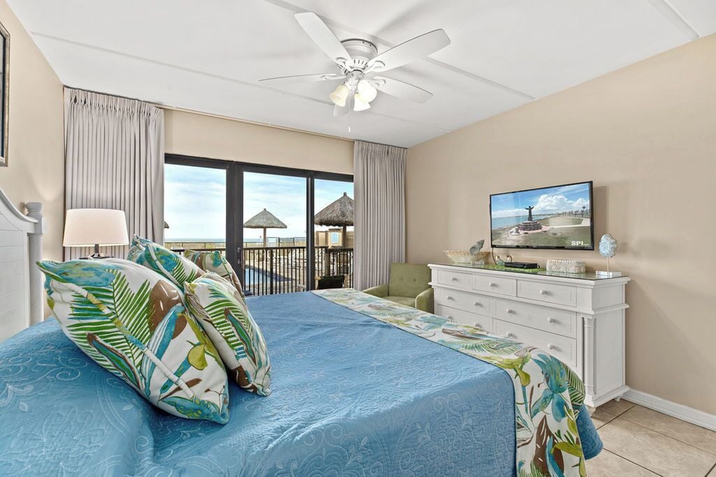 Hotelkamer met een groot bed, een plafondventilator, een televisie en uitzicht op het strand.