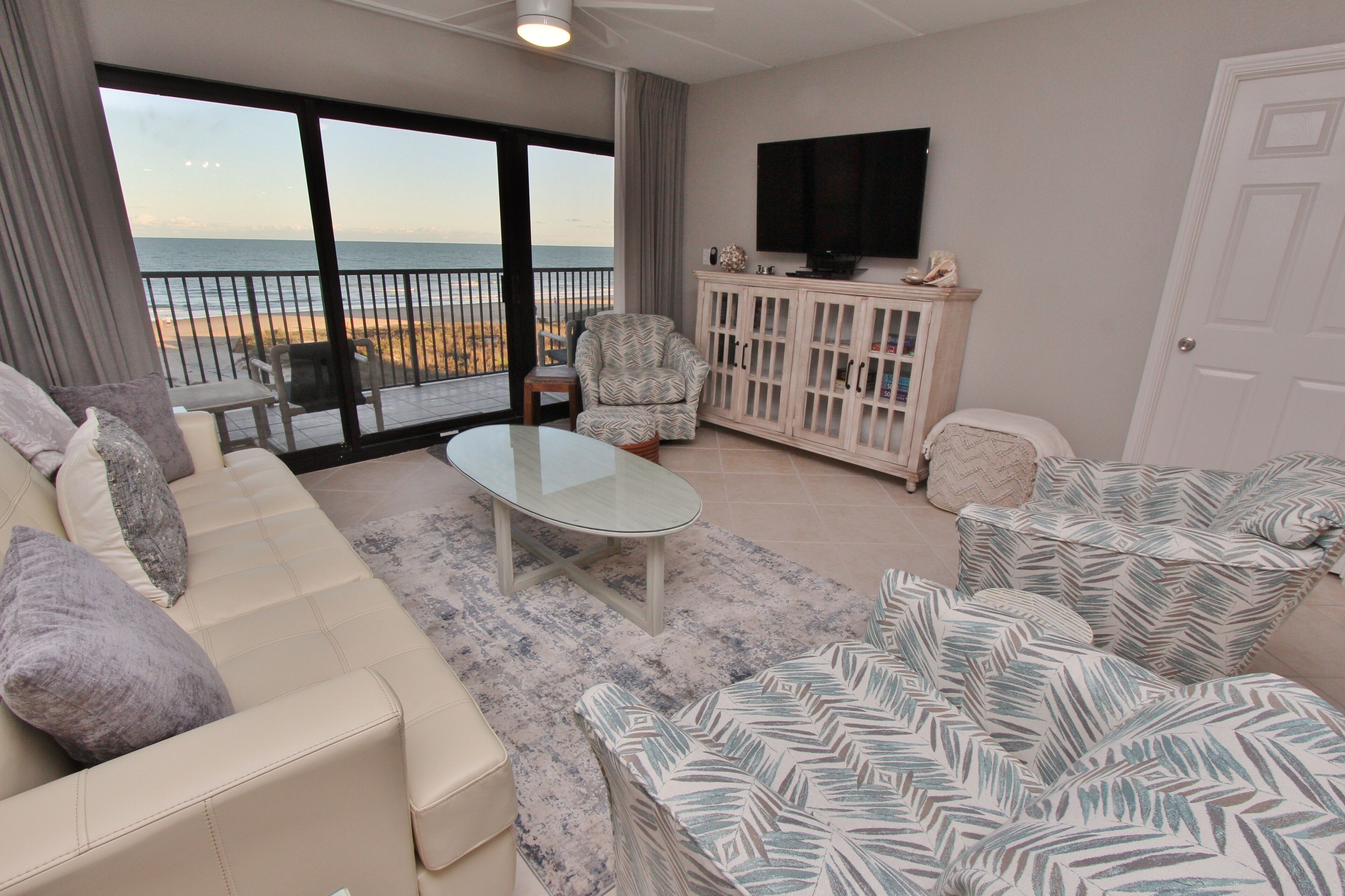 Un salon comprenant un canapé, un fauteuil et une table basse en verre. Un téléviseur est fixé au mur et de grandes fenêtres offrent une vue sur la plage.