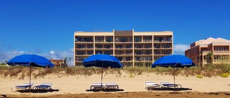 A beachfront hotel with multiple balconies, beach umbrellas, and lounge chairs.