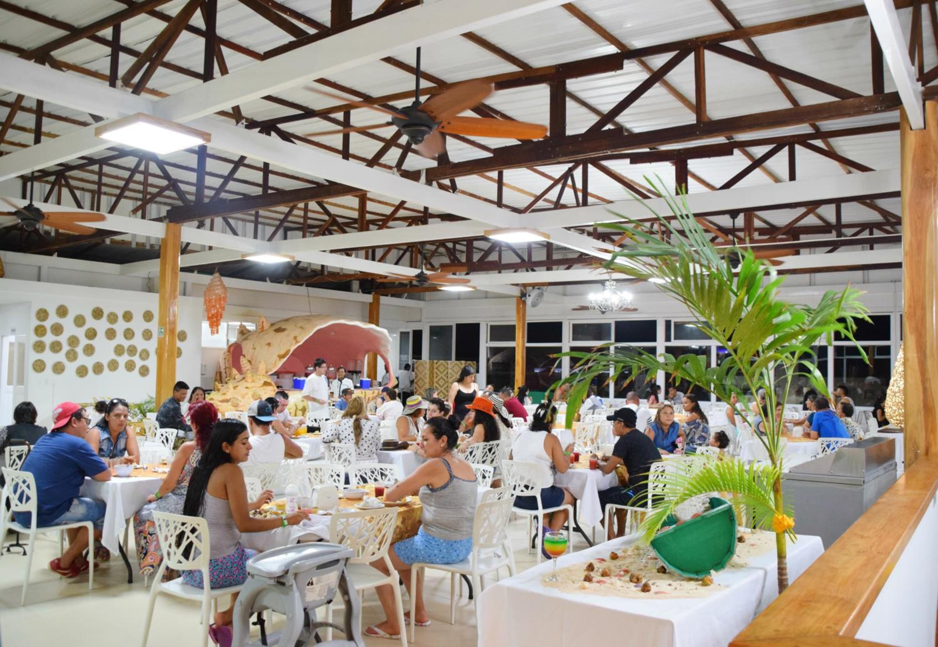 A spacious indoor dining area with white tables and chairs, ceiling fans, and decorative elements.