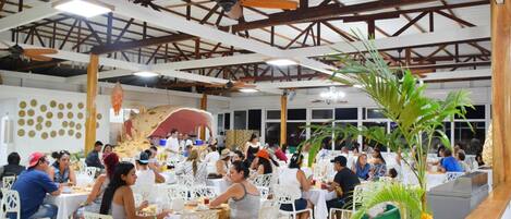 A spacious indoor dining area with white tables and chairs, ceiling fans, and decorative elements.