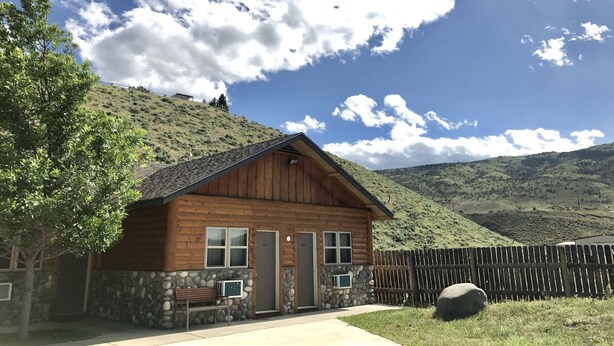 A wooden cabin with a stone foundation, a porch, and a view of a hilly landscape. - Cody Holiday Lodge (Cody)