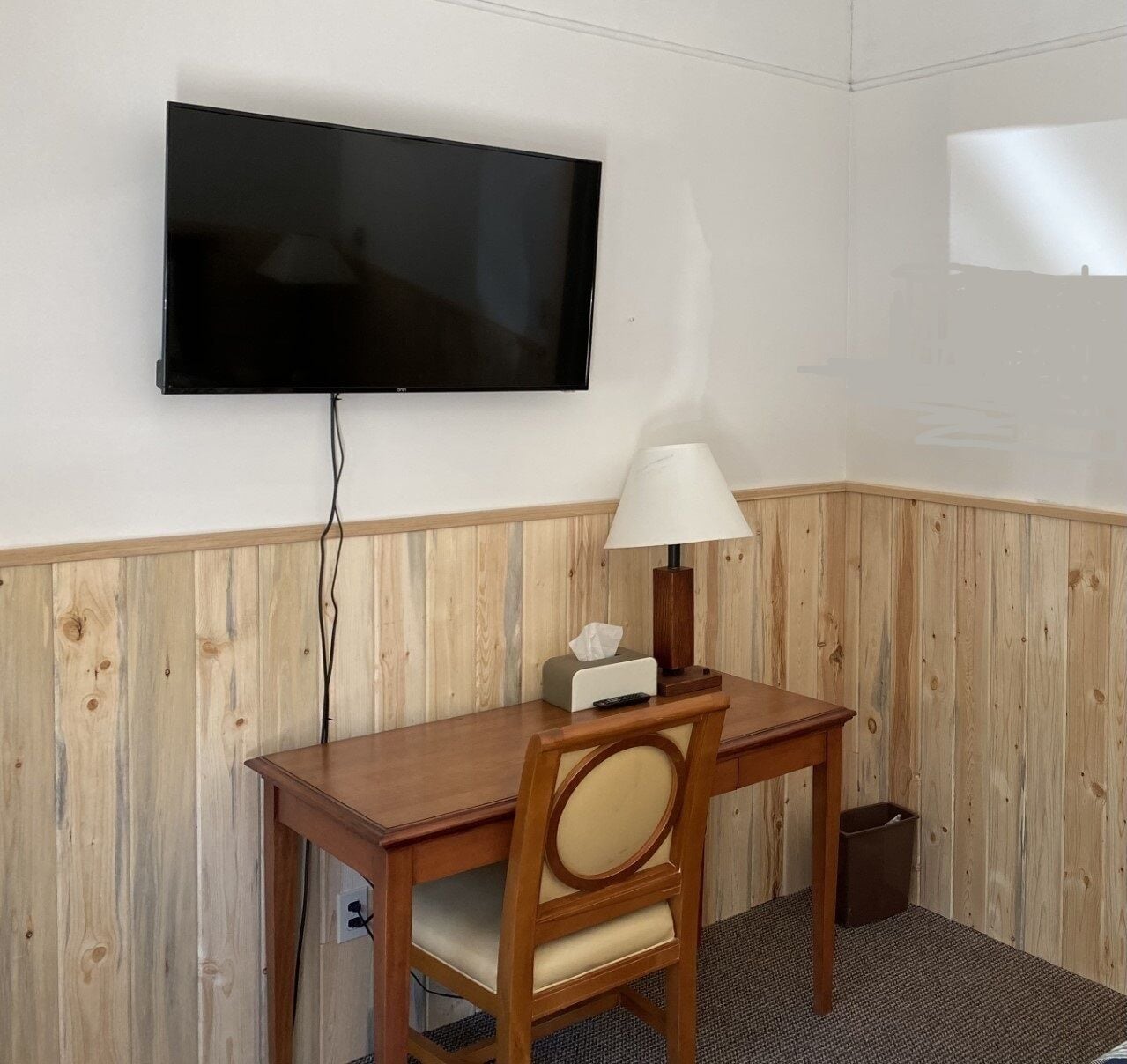 A hotel room with a wooden paneled wall, a flat-screen TV mounted above a wooden desk, a lamp, a tissue box, and a trash bin.