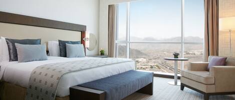 Ein Hotelzimmer mit einem großen Bett, einer Bank, einem Sessel, einem kleinen Tisch und Blick auf eine Stadt durch große Fenster.