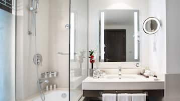 Une salle de bain moderne avec une douche à parois de verre, un grand miroir et un meuble-lavabo blanc où sont rangés les articles de toilette.